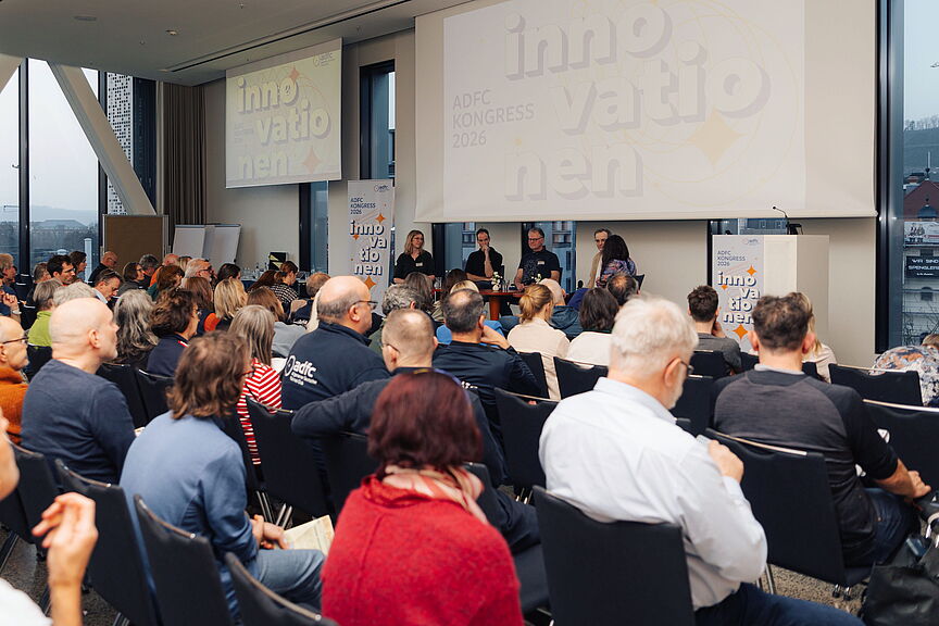 ADFC-Kongress 2026 in Würzburg Blick über die Köpfe zahlreicher sitzender Zuschauer im Saal auf eine Bühne. Dort diskutieren vier Personen bei einer Podiumsdiskussion. Im Hintergrund zeigen Leinwände den Schriftzug ADFC Kongress 2026 Innovationen.